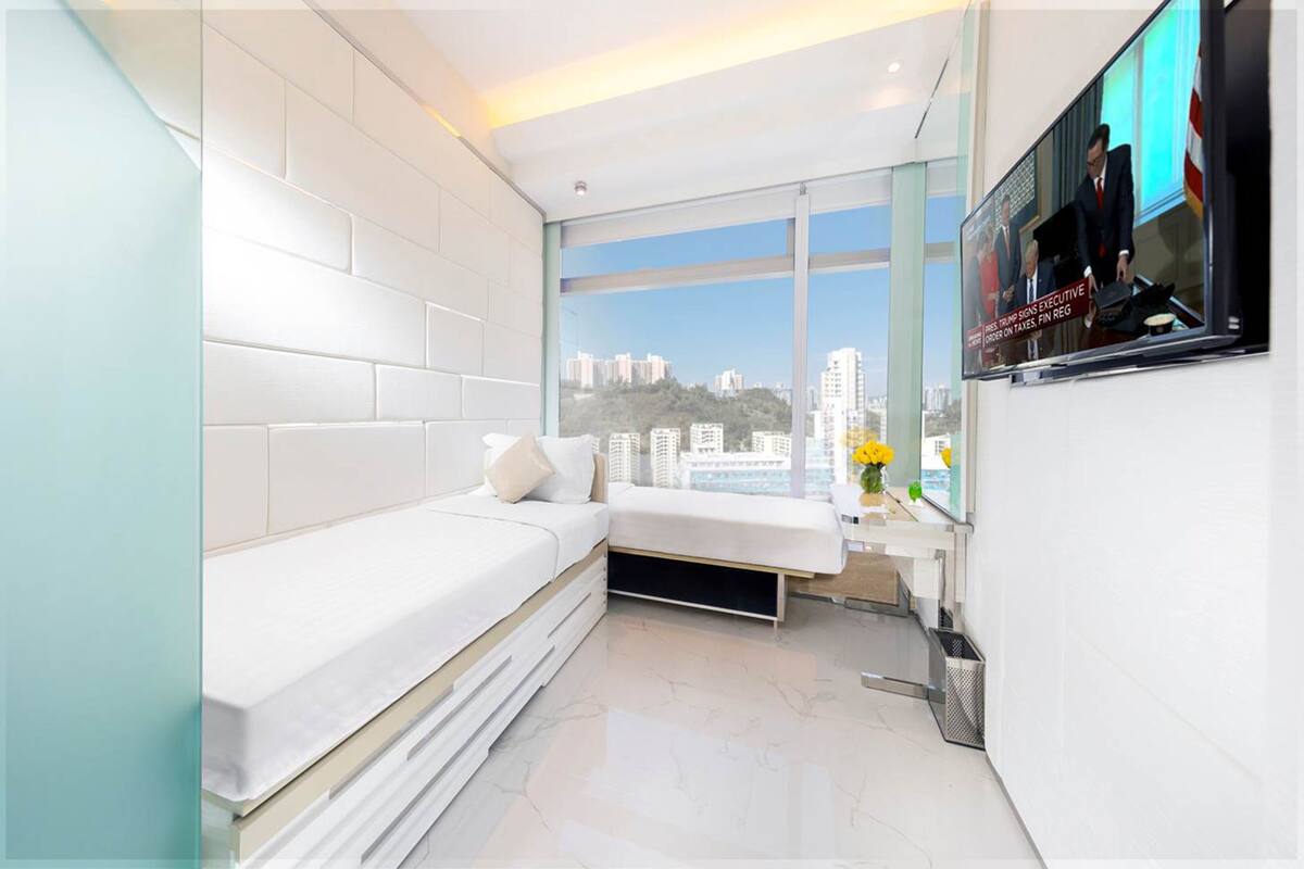A modern hotel room with a white bed, a small seating area, a large window with a city view, and a TV mounted on the wall.