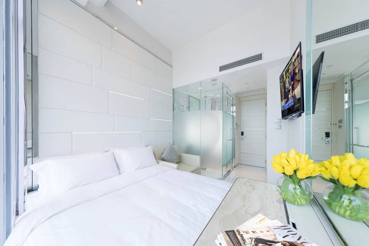 A modern hotel room with a large bed, a glass shower enclosure, and a flat-screen TV mounted on the wall.