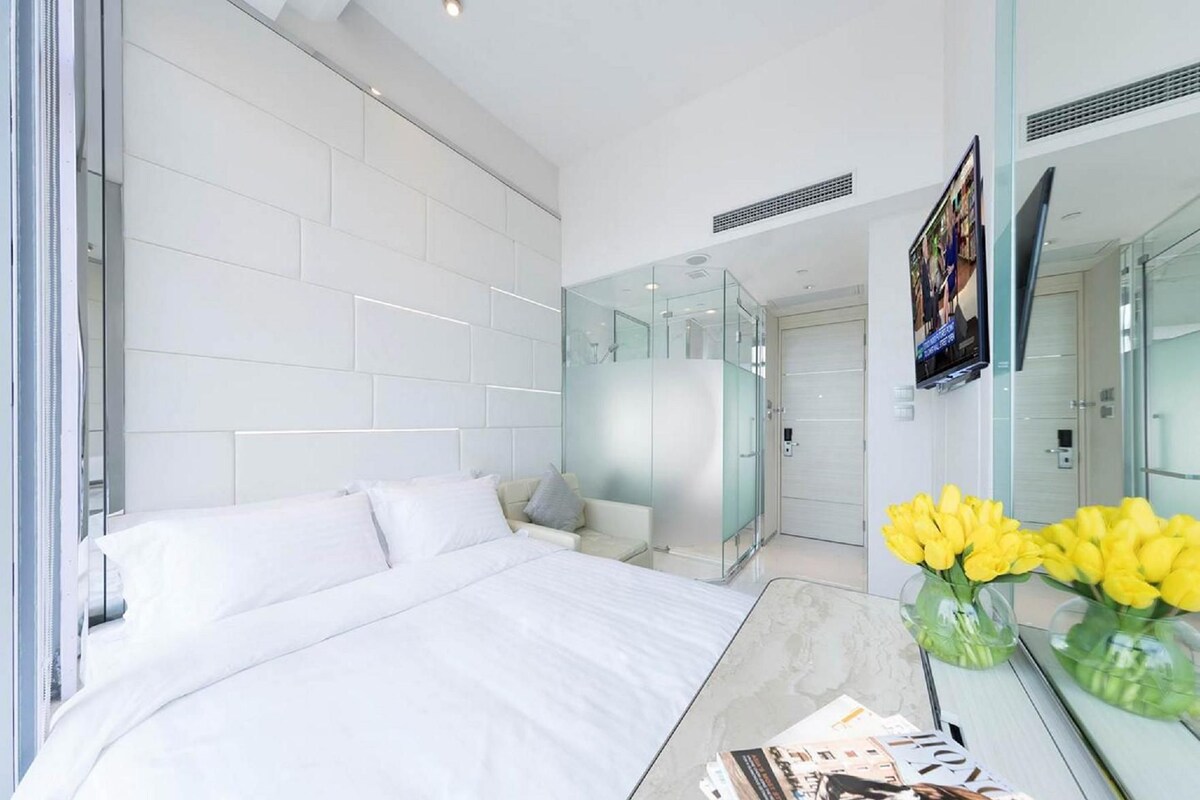 Ein modernes Hotelzimmer mit einem großen Bett, einer Glaskabine, einem Flachbildfernseher und einer Vase mit gelben Blumen.