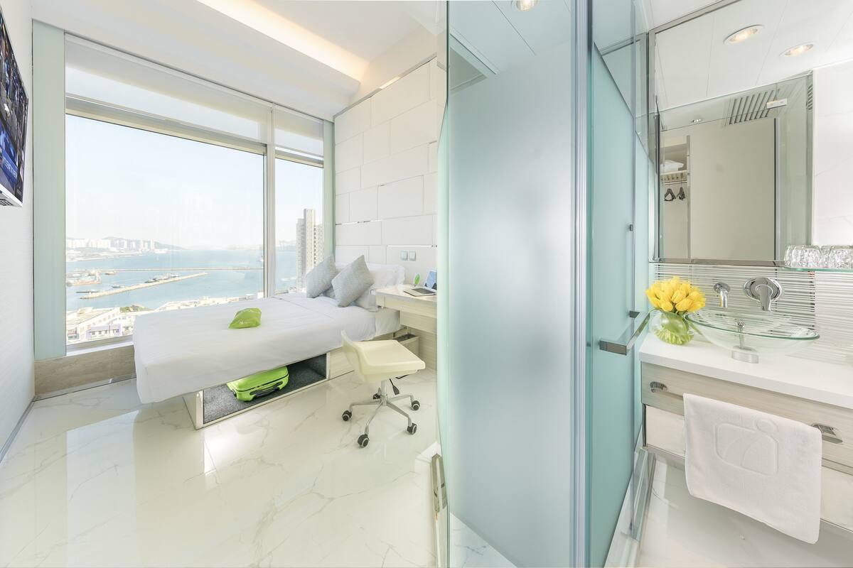 A modern hotel room with a large window, a bed, a desk, a glass shower enclosure, and a bathroom with a large mirror and sink.