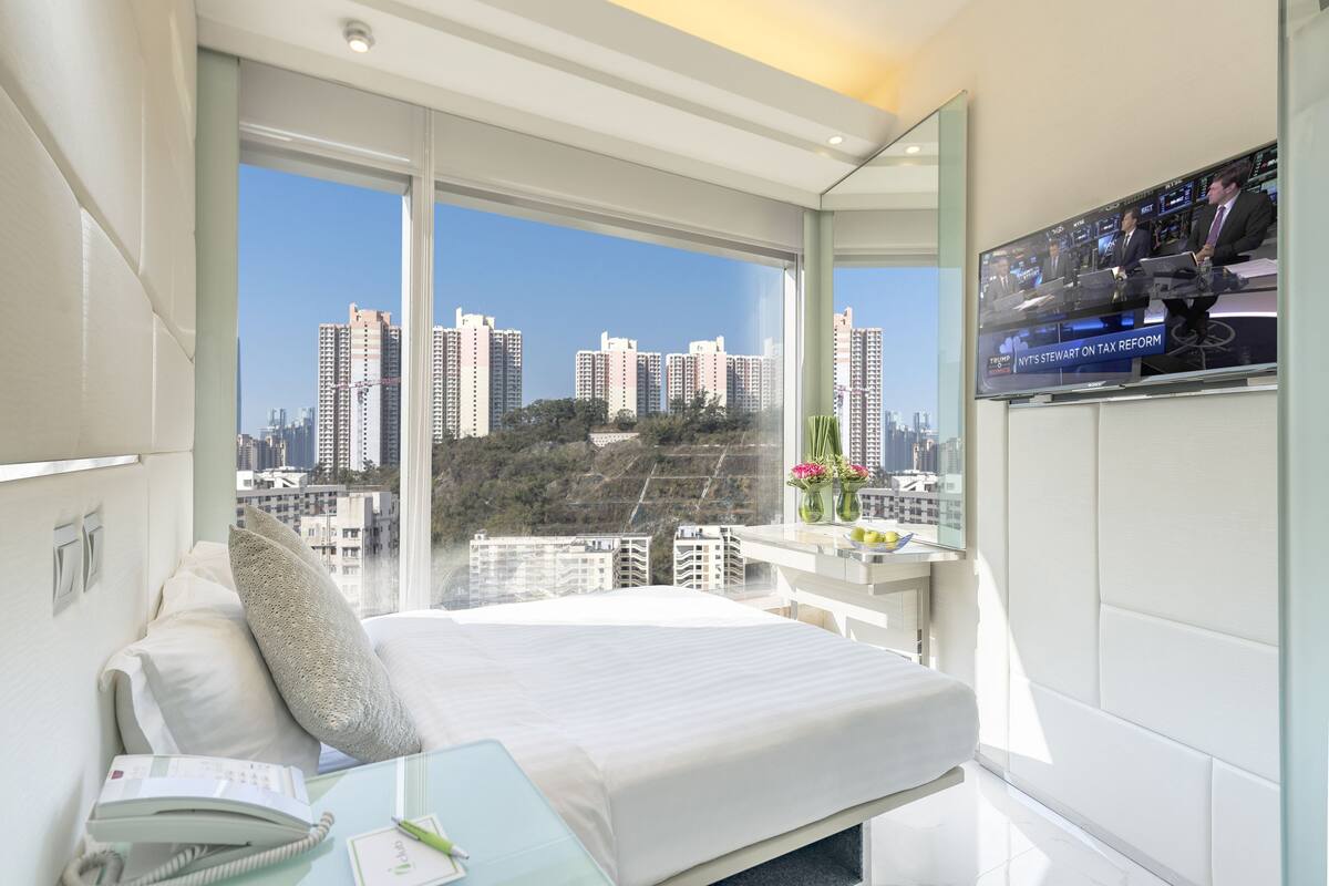 A modern hotel room with a large bed, a desk with a chair, a flat-screen TV, and a city view.