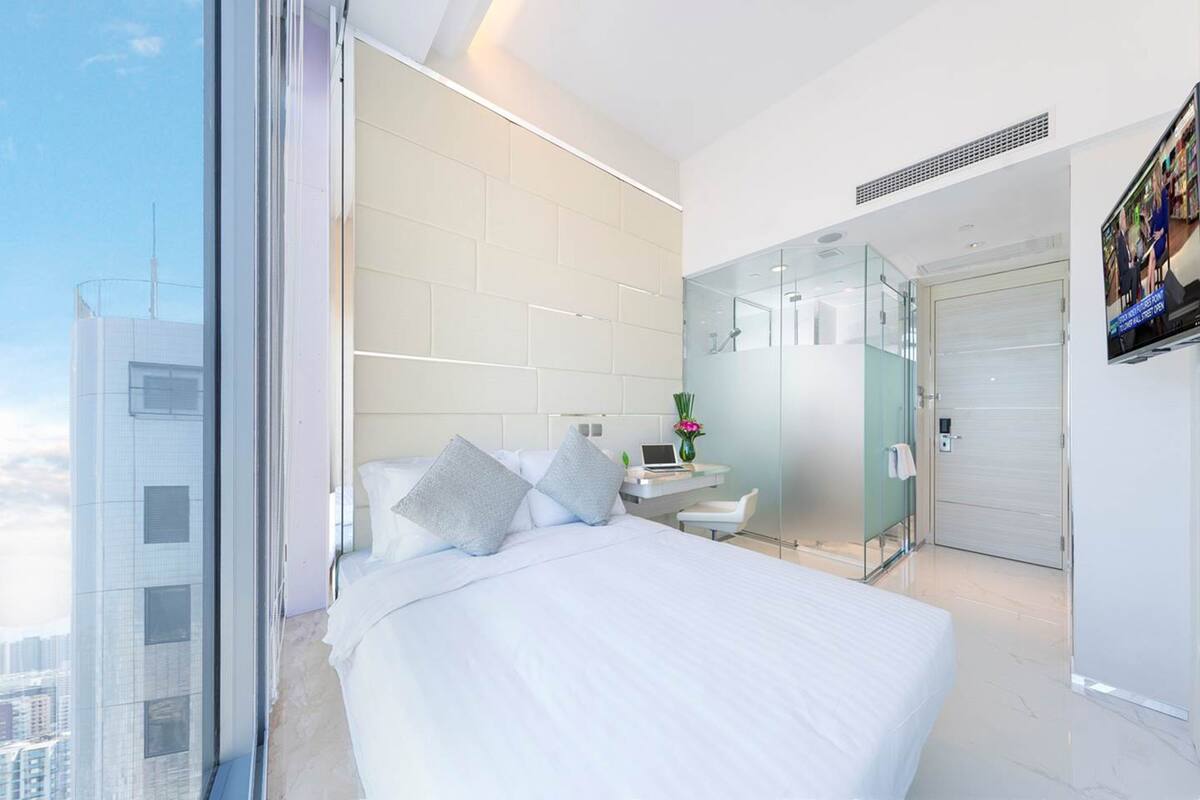 A modern hotel room with a large bed, a small desk, a glass shower enclosure, and a city view.