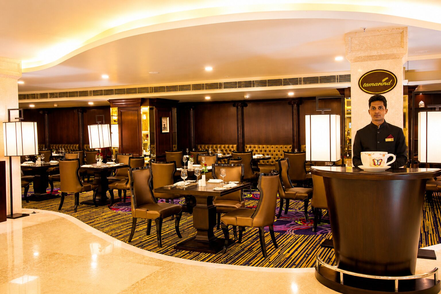 Eine Hotellobby mit mehreren Essplätzen, Tischen, Stühlen, einer Rezeption und einer großen Säule mit einem Logo.