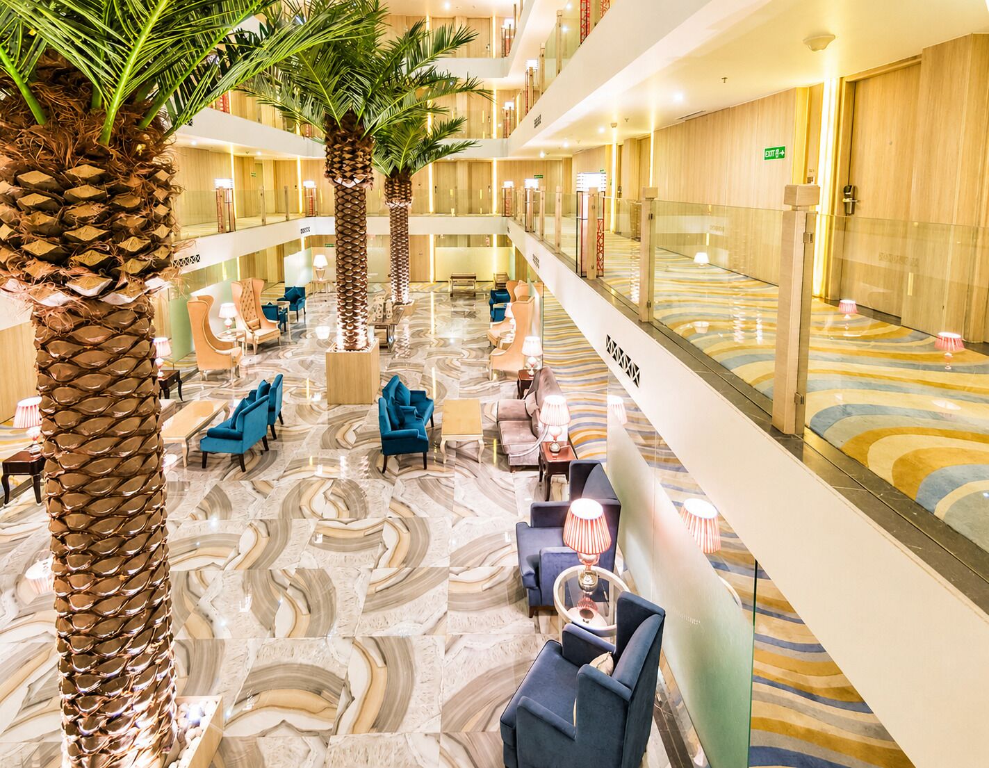 Un vestíbulo de hotel con suelo de mármol, zonas de estar, palmeras y una barandilla de cristal que da al nivel inferior.
