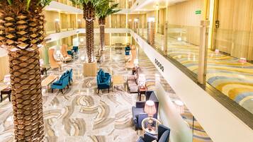 Un vestíbulo de hotel con suelo de mármol, zonas de estar, palmeras y una barandilla de cristal que da al nivel inferior.