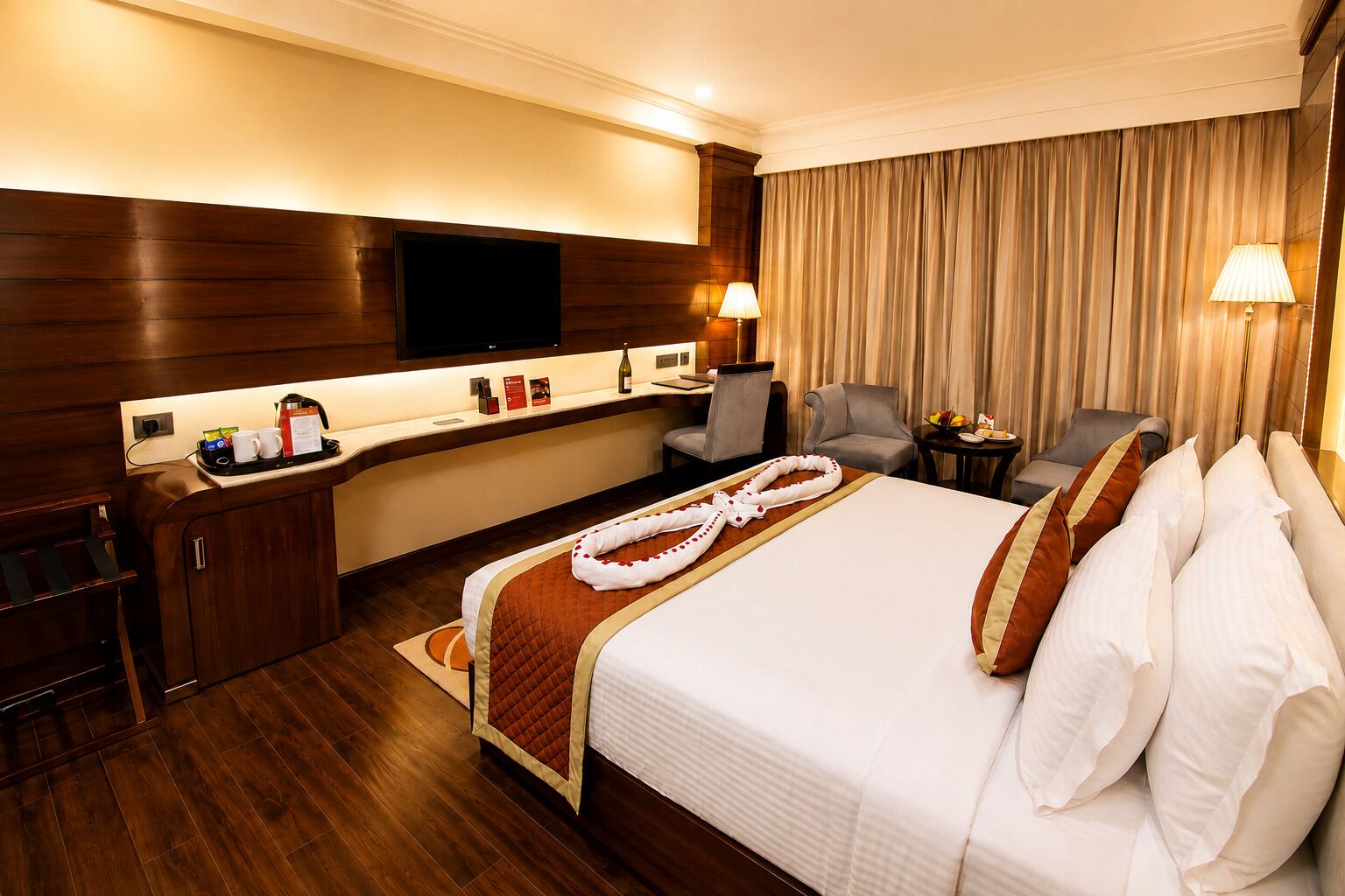 A hotel room with a large bed, a desk with a flat-screen TV, a minibar, and a seating area with a chair and a small table.