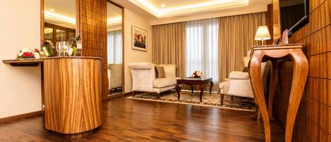A hotel room with a wooden floor, a sofa set, a coffee table, a TV mounted on the wall, and a wooden sideboard with a lamp.