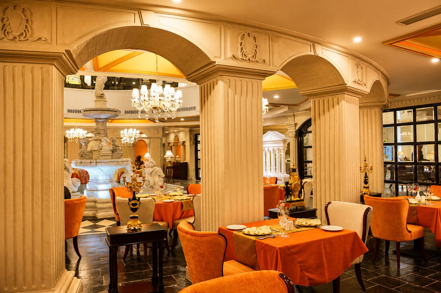 A dining area with tables set for a meal, featuring a fountain and classical architecture.