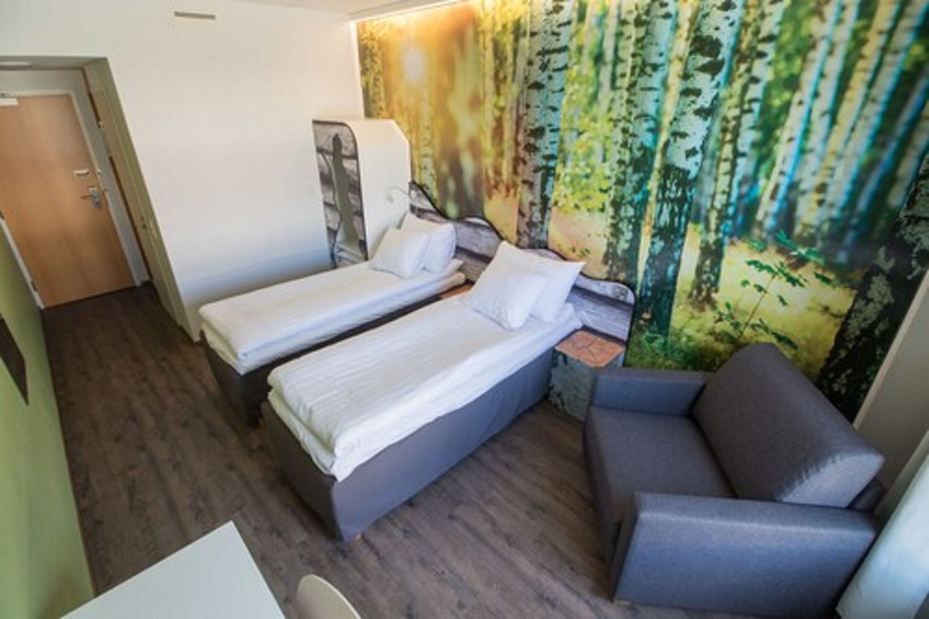 A compact room with two single beds, a gray sofa, and a wall mural of a forest.