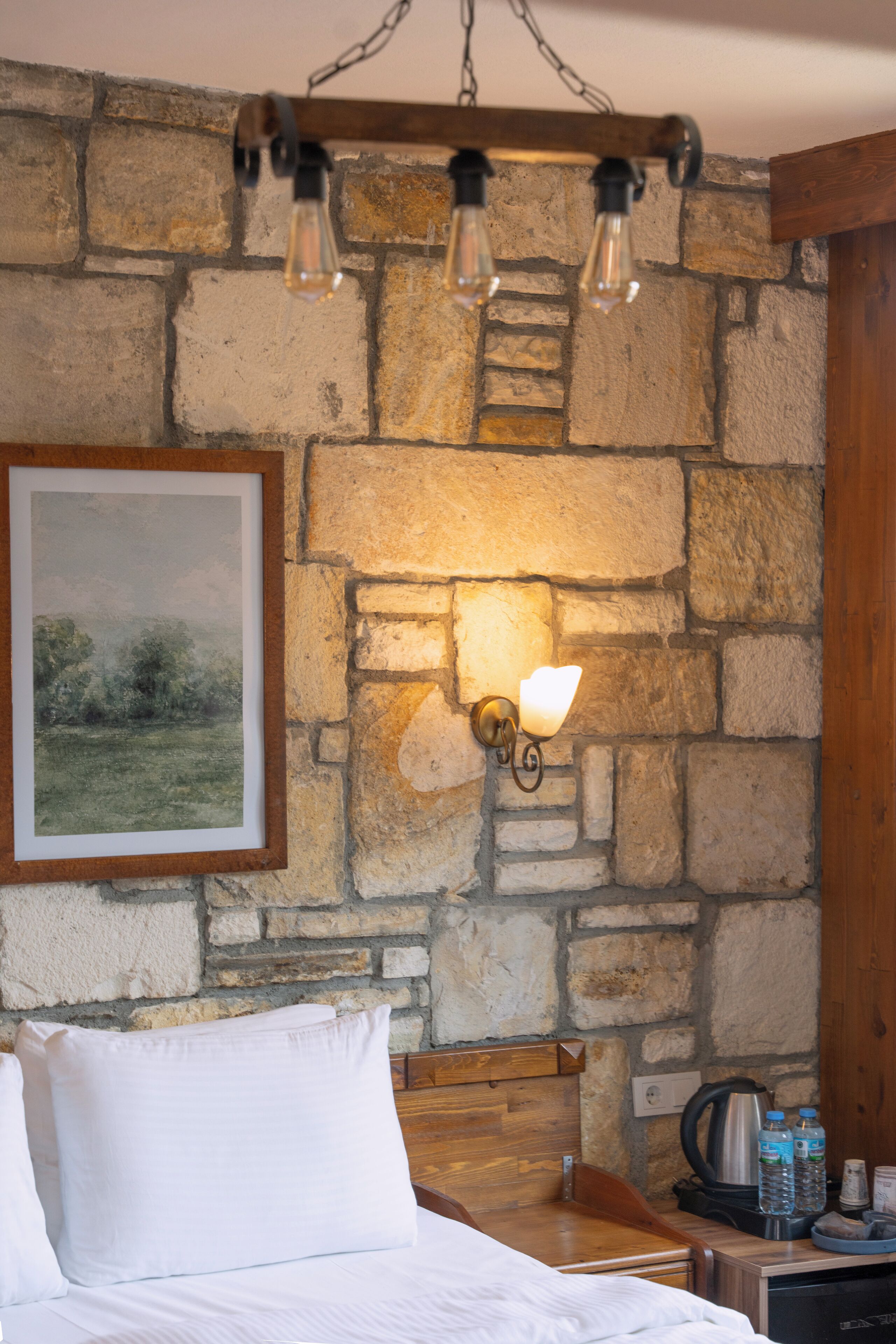 Una habitación con pared de piedra, una cama con ropa de cama blanca, un cuadro enmarcado, una mesita de noche con tetera y una lámpara de pared.