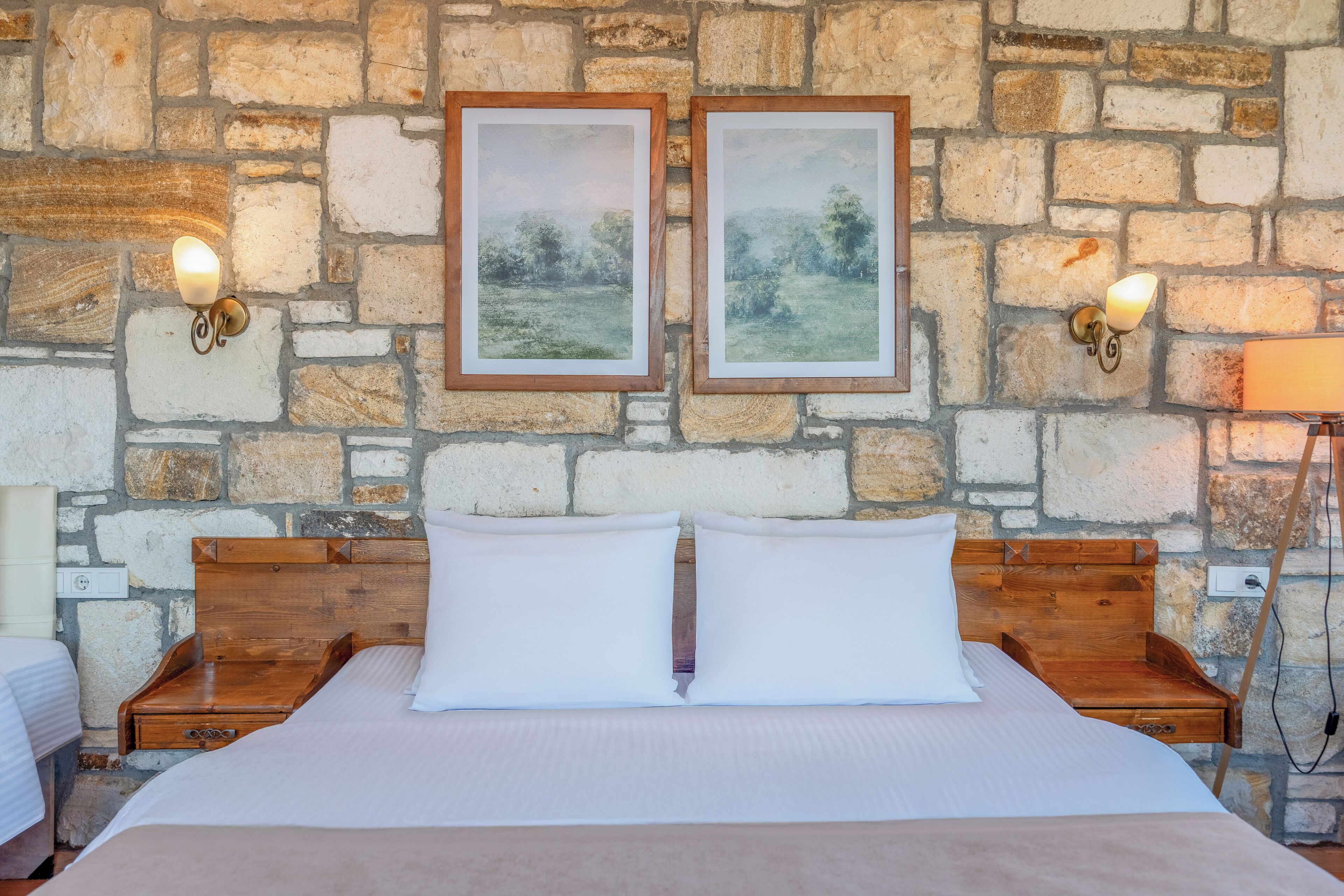 Un lit double avec une tête de lit en bois et deux oreillers blancs, placé contre un mur de pierre orné de deux tableaux encadrés et de lampes fixées au mur.