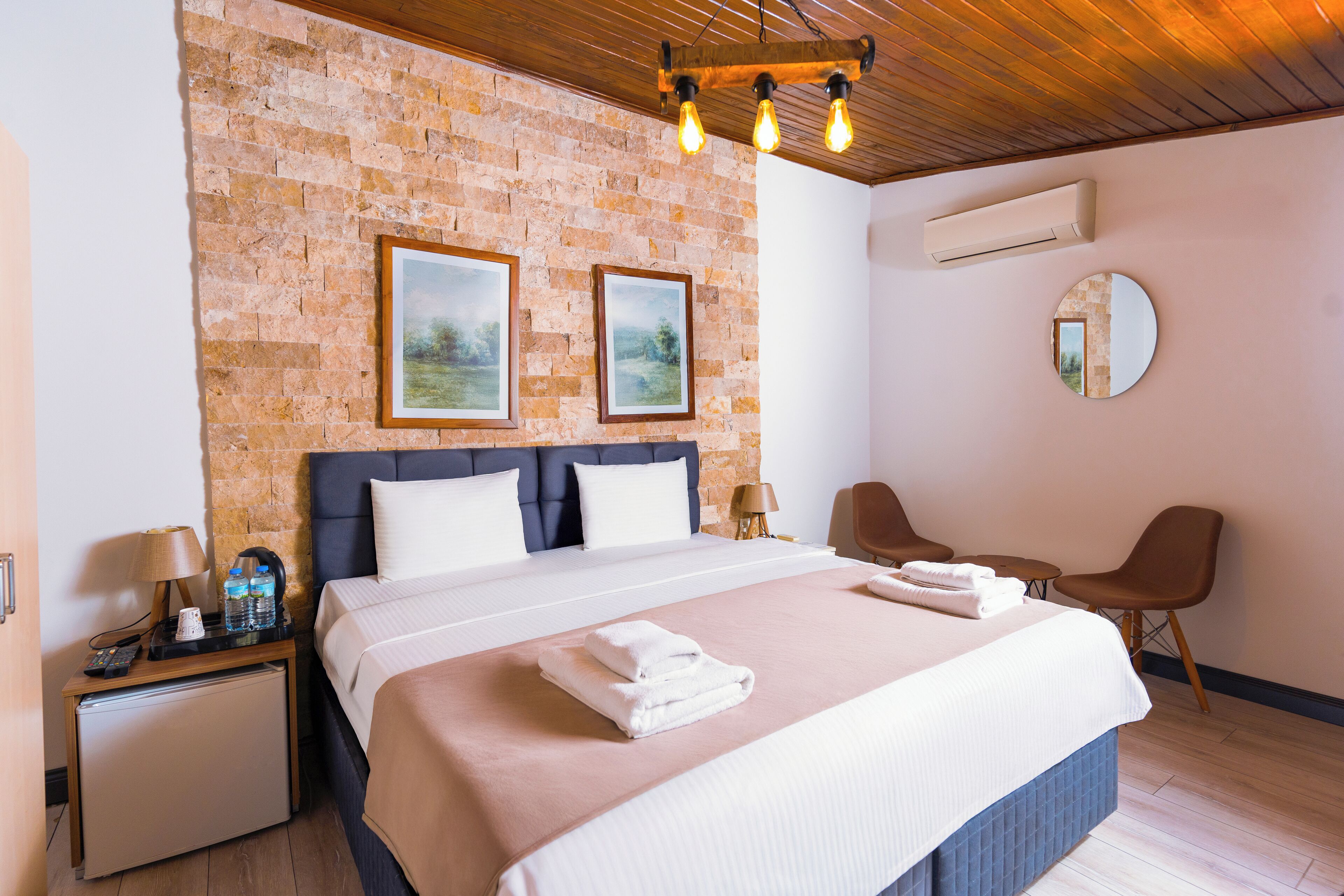 A neatly arranged hotel room with a brick wall, a bed with a blue headboard, a round mirror, and a small table with a chair.