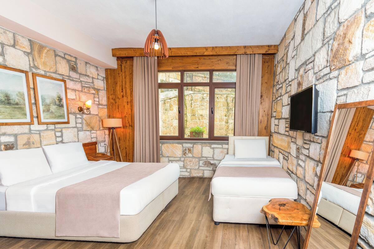 Una habitación con dos camas, paredes de piedra, vigas de madera, televisión y una ventana con cortinas.