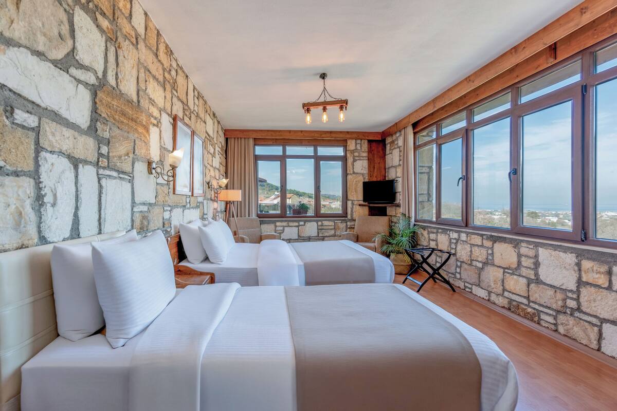Habitación de hotel con paredes de piedra, dos camas, televisión y ventanas amplias con vistas.