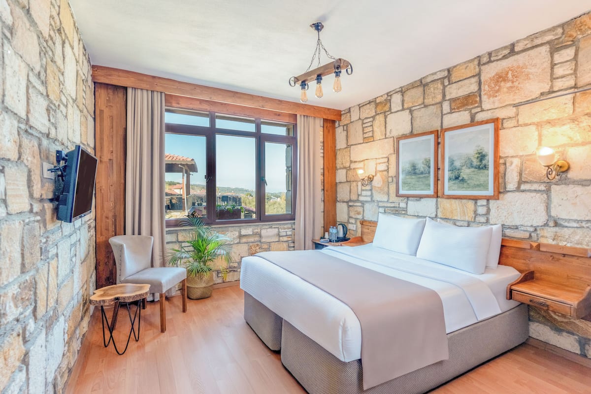 Un dormitorio con pared de piedra, una cama grande, un televisor, una silla, una mesita y una ventana con vistas.