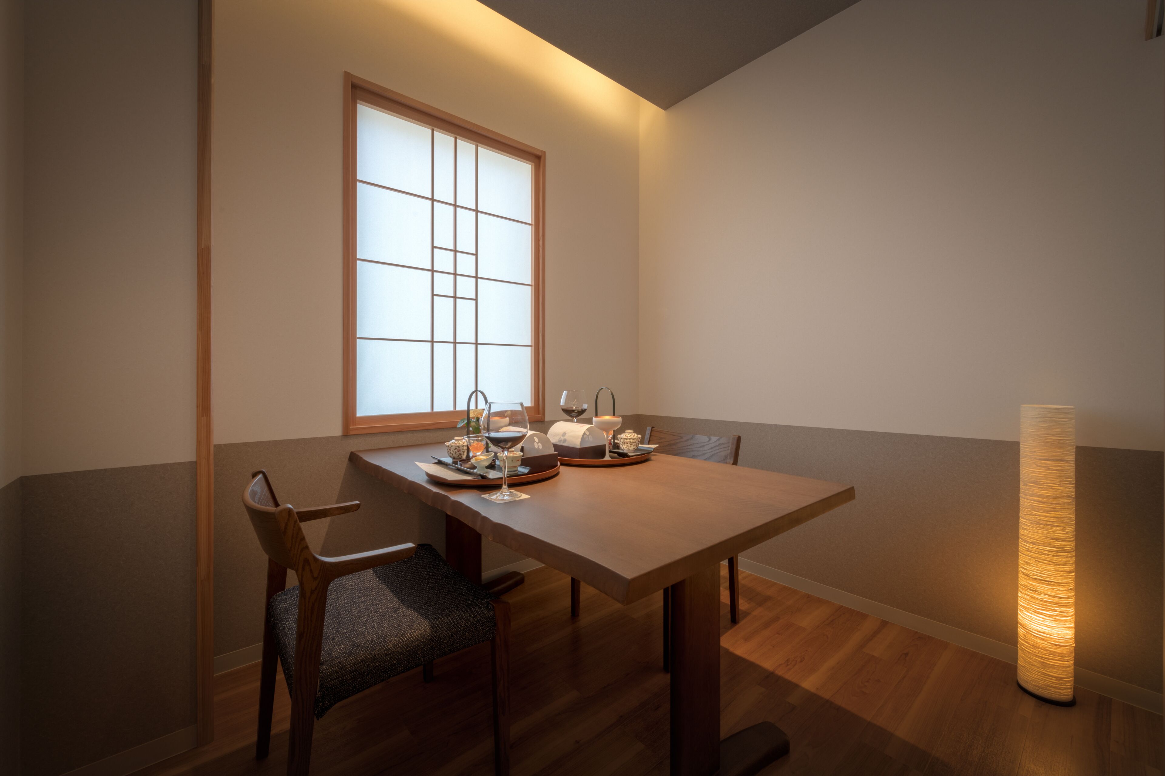 A dining area with a wooden table, chairs, a window with a grid pattern, and a tall, cylindrical light fixture.