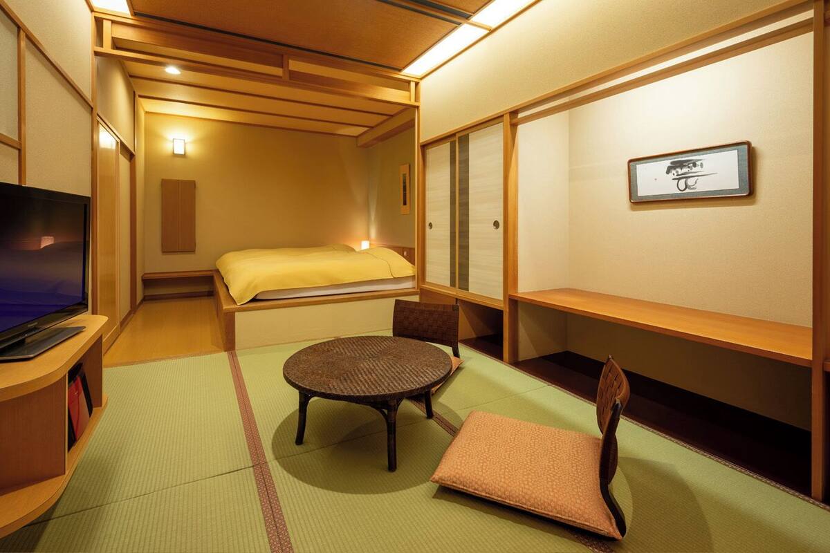 A traditional Japanese room with tatami flooring, a bed, a television, and a small round table with a chair.