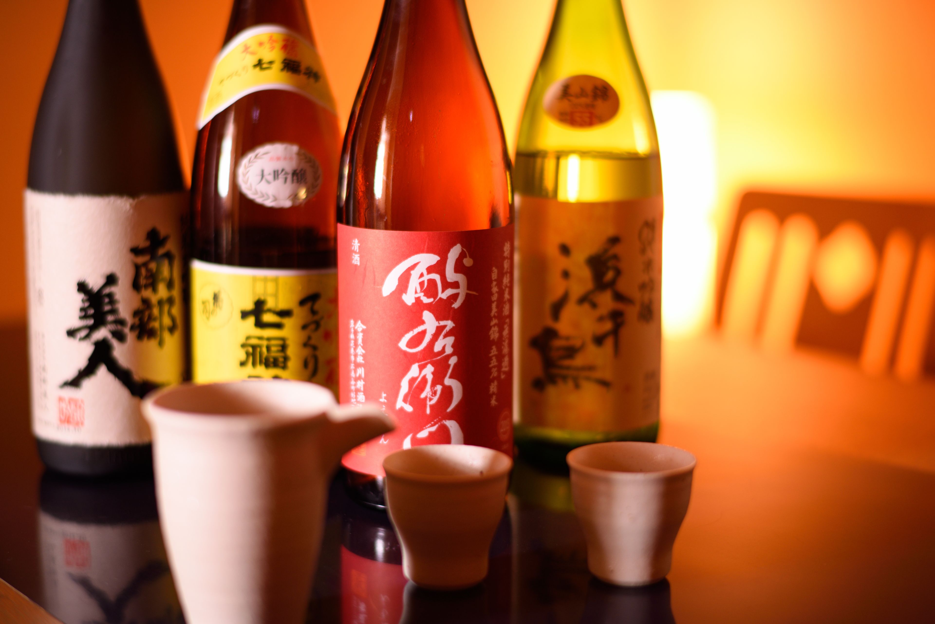 A selection of sake bottles with labels, some with Japanese characters, and small ceramic cups in front.
