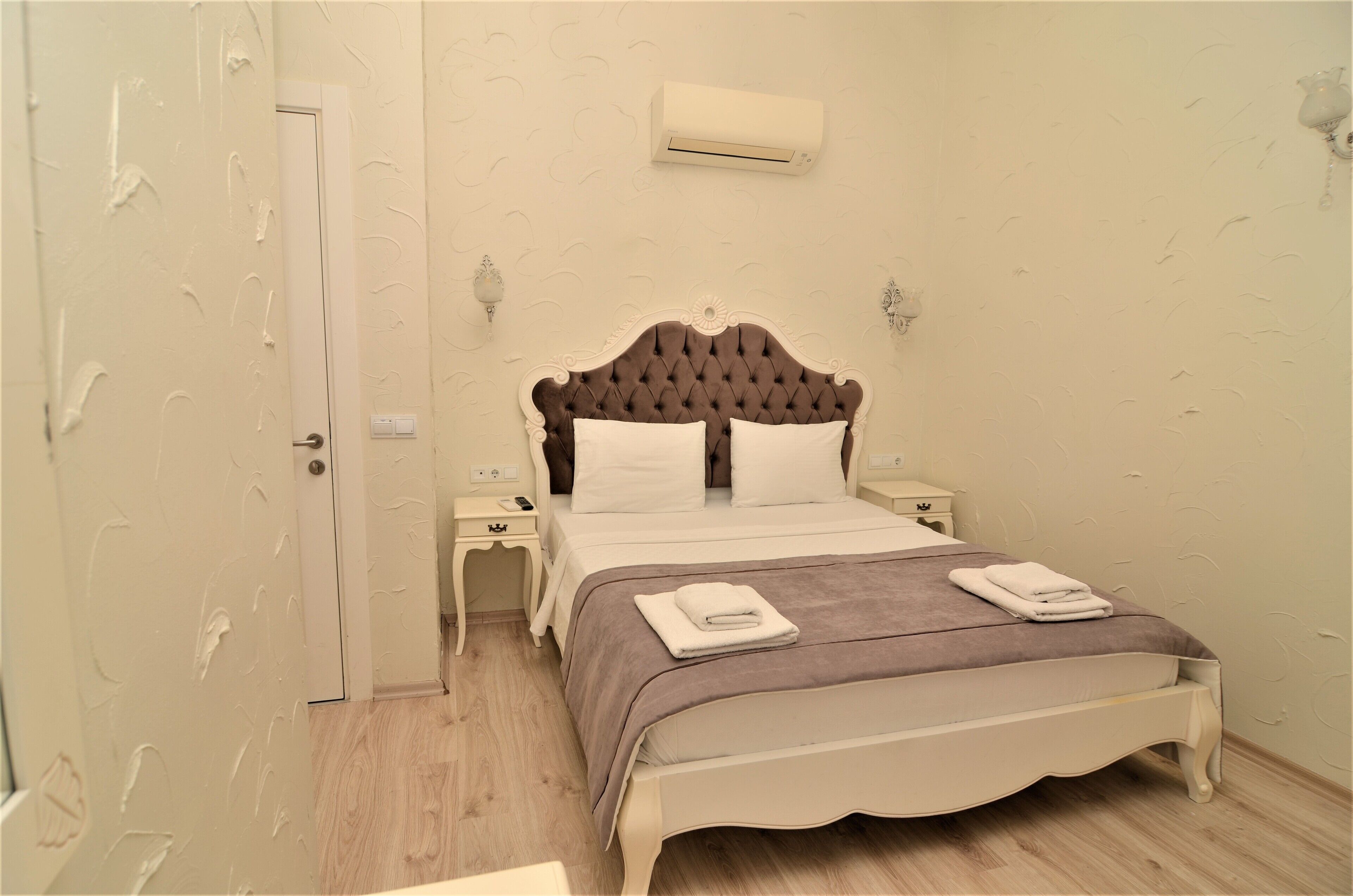 Une chambre à coucher avec une tête de lit capitonnée, deux tables de chevet, un lit recouvert d’une couverture marron et un appareil de climatisation fixé au mur.