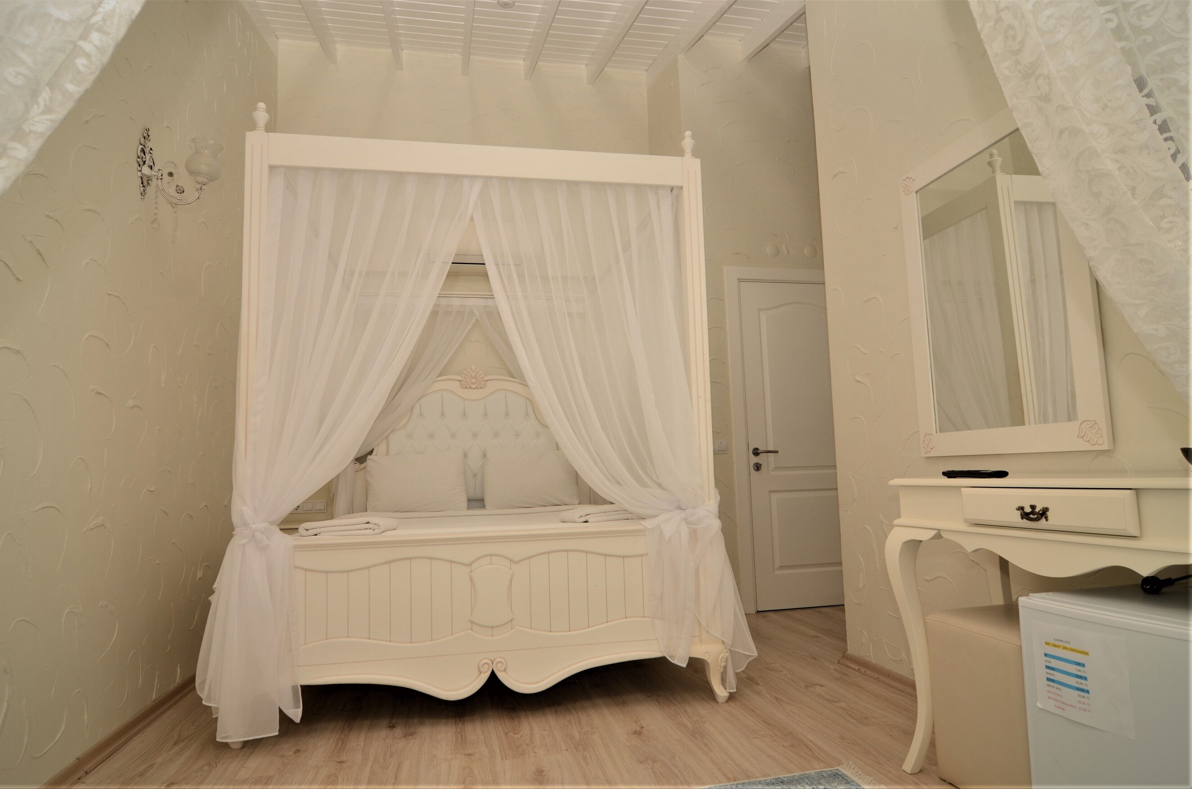 A bedroom with a four-poster bed, a bedside table, a mirror, and a door.