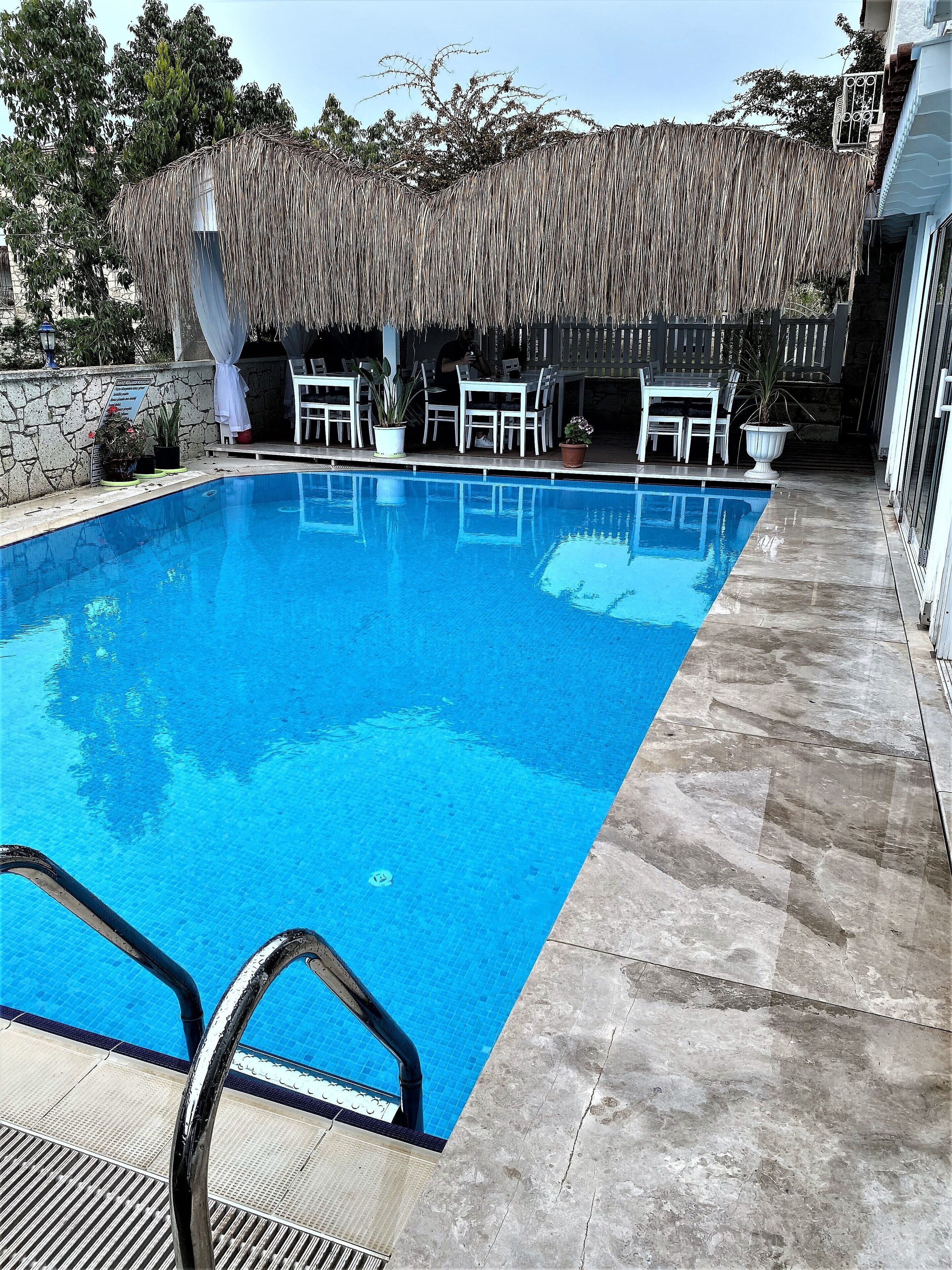 Una piscina rectangular con agua azul cristalina, rodeada por un muro de piedra y una estructura de techo de paja.