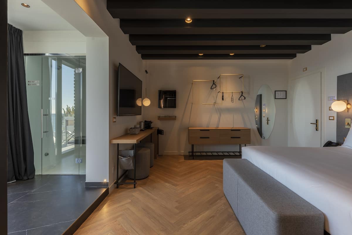A modern hotel room with a large bed, a desk with a chair, a wardrobe, a mirror, and a glass door leading to a balcony.