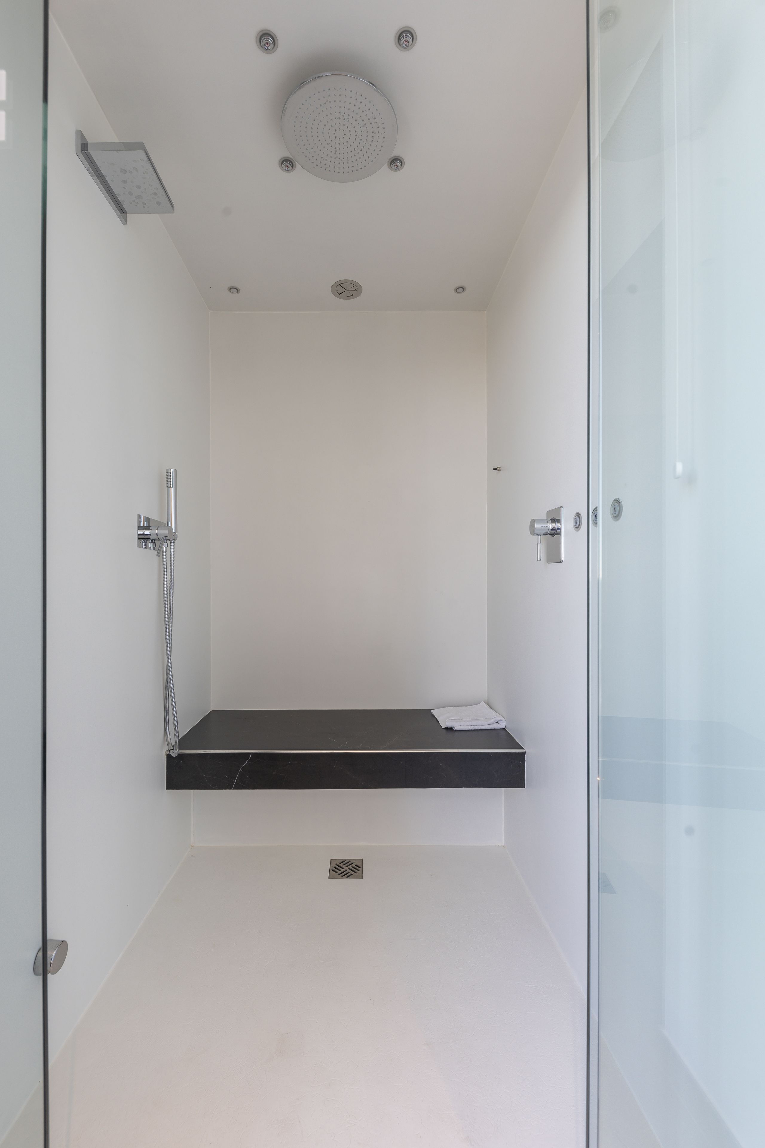Une salle de douche moderne dotée d’une porte en verre, d’un banc noir et d’une pomme de douche fixée au plafond.