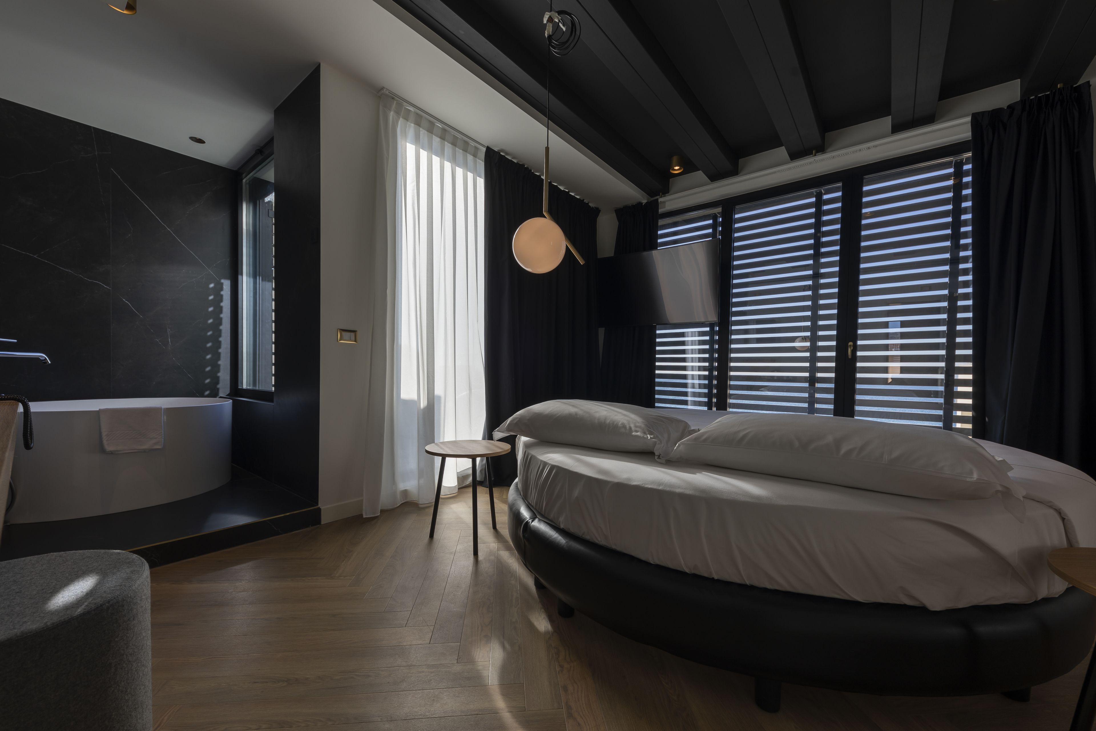 Ein modernes Schlafzimmer mit einem großen Bett, einem kleinen runden Tisch, einer Badewanne und einem großen Fenster mit Jalousien.