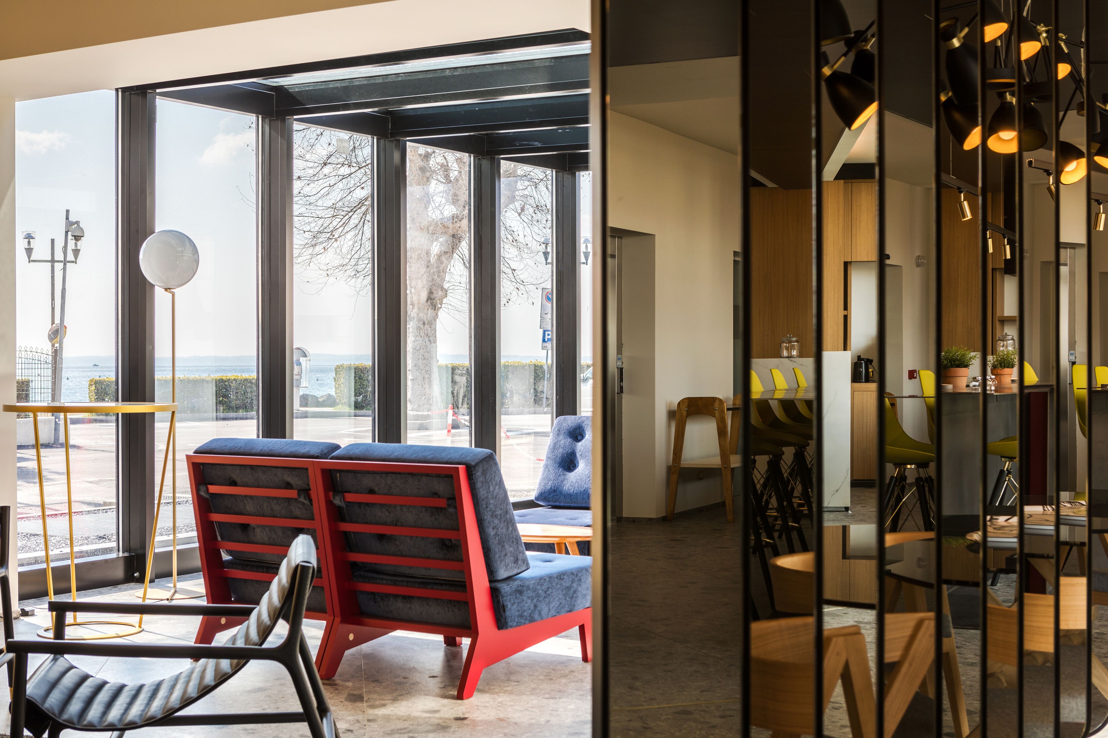 A modern interior with large windows, a seating area with red and blue chairs, and a kitchen visible through a glass partition.