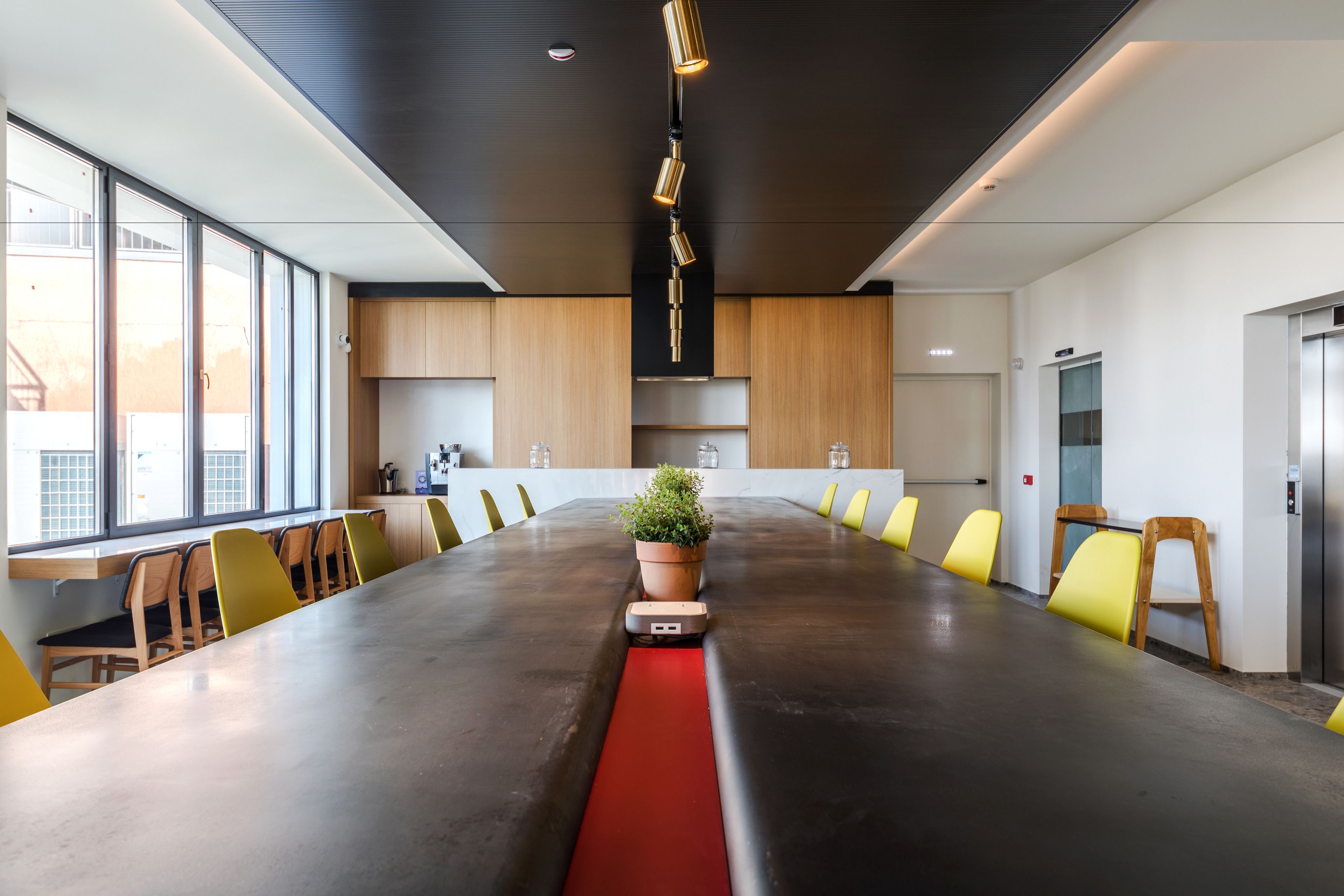 A modern conference room with a long table, yellow chairs, a potted plant, and a red strip on the table.