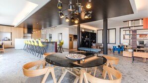 Een modern interieur met een ronde glazen tafel, houten stoelen, een bar met gele stoelen en een plafond met hanglampen.
