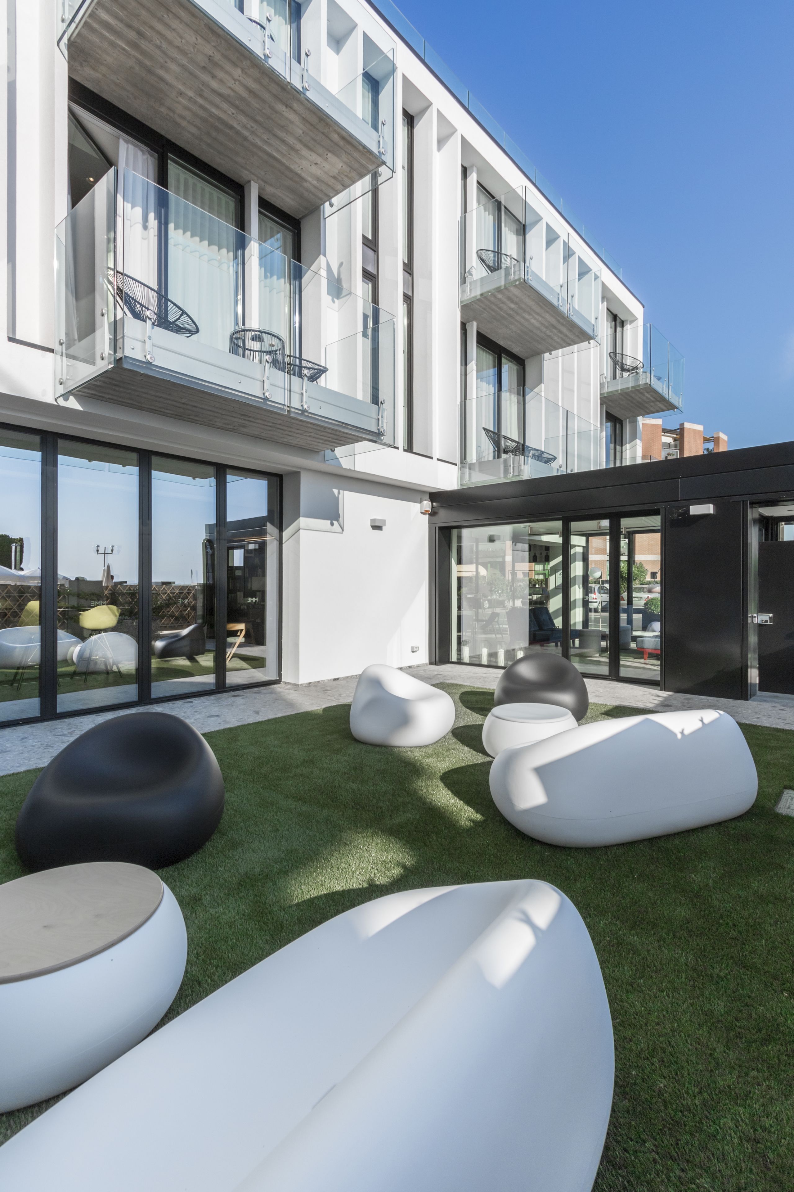 Un edificio moderno con amplios ventanales, mobiliario exterior con cojines blancos y negros, y un césped bien cuidado.