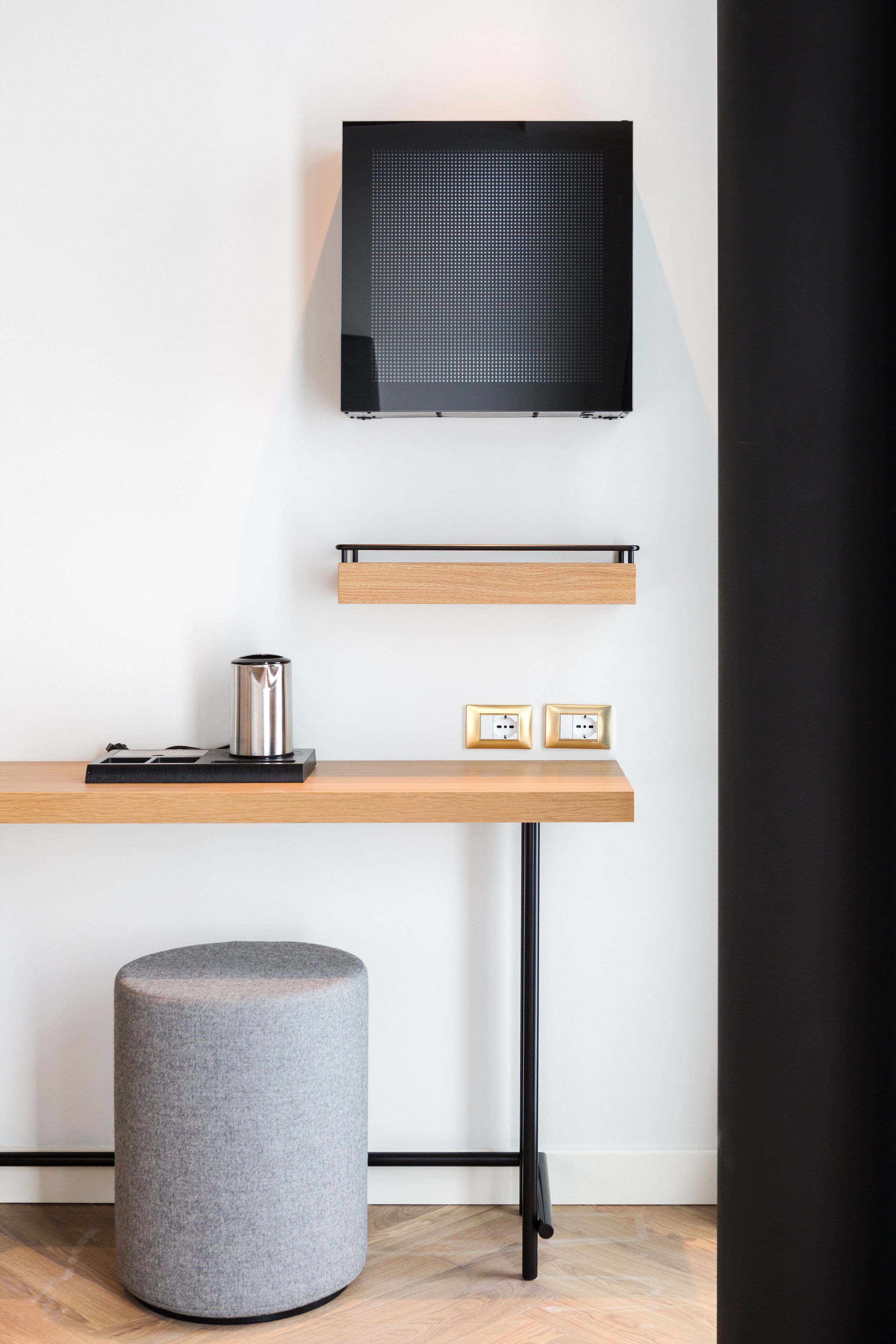Una habitación moderna con un escritorio de madera, un taburete gris, televisor montado y enchufes.