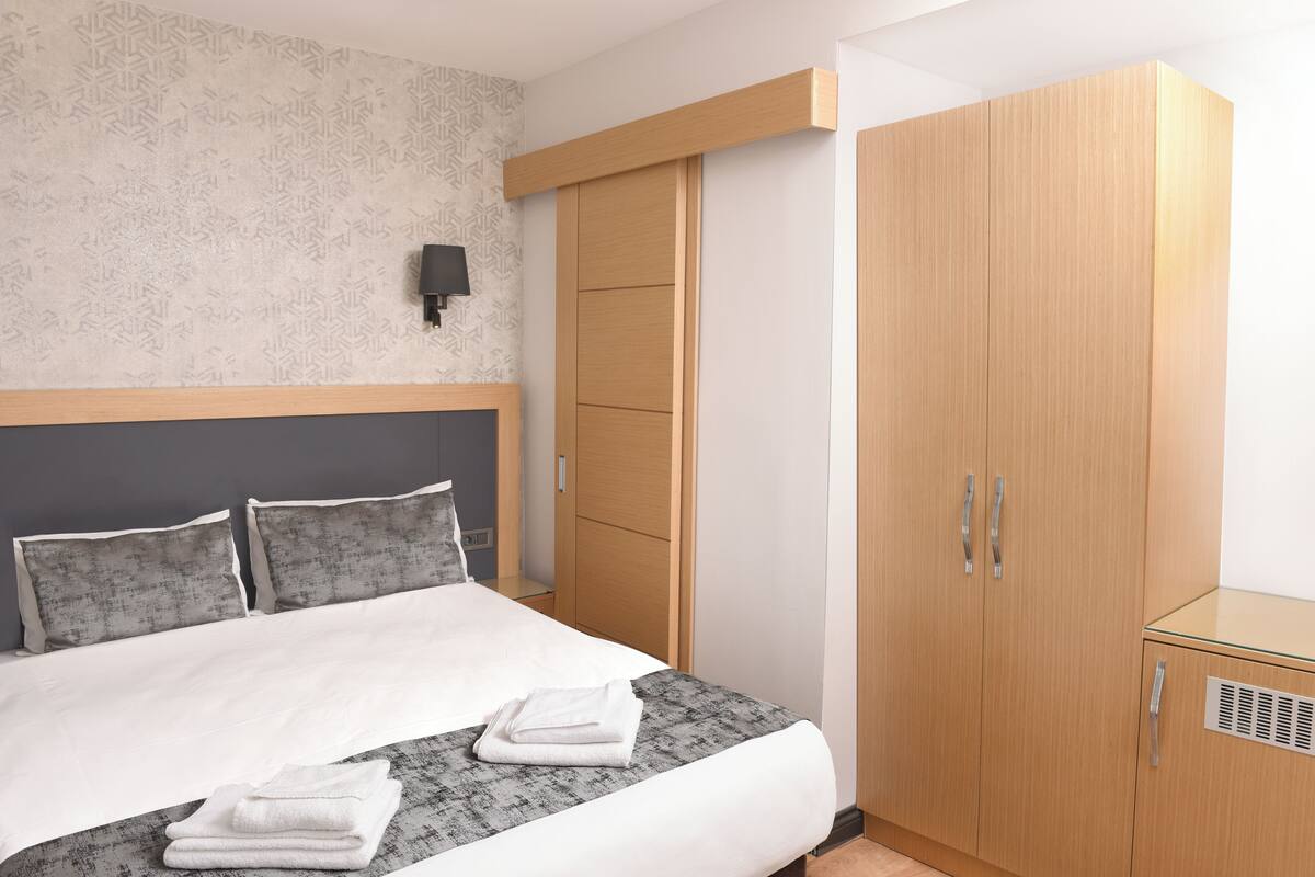 A neatly made bed with a grey and white patterned bedspread, a wooden wardrobe with two doors, and a wall-mounted light fixture.