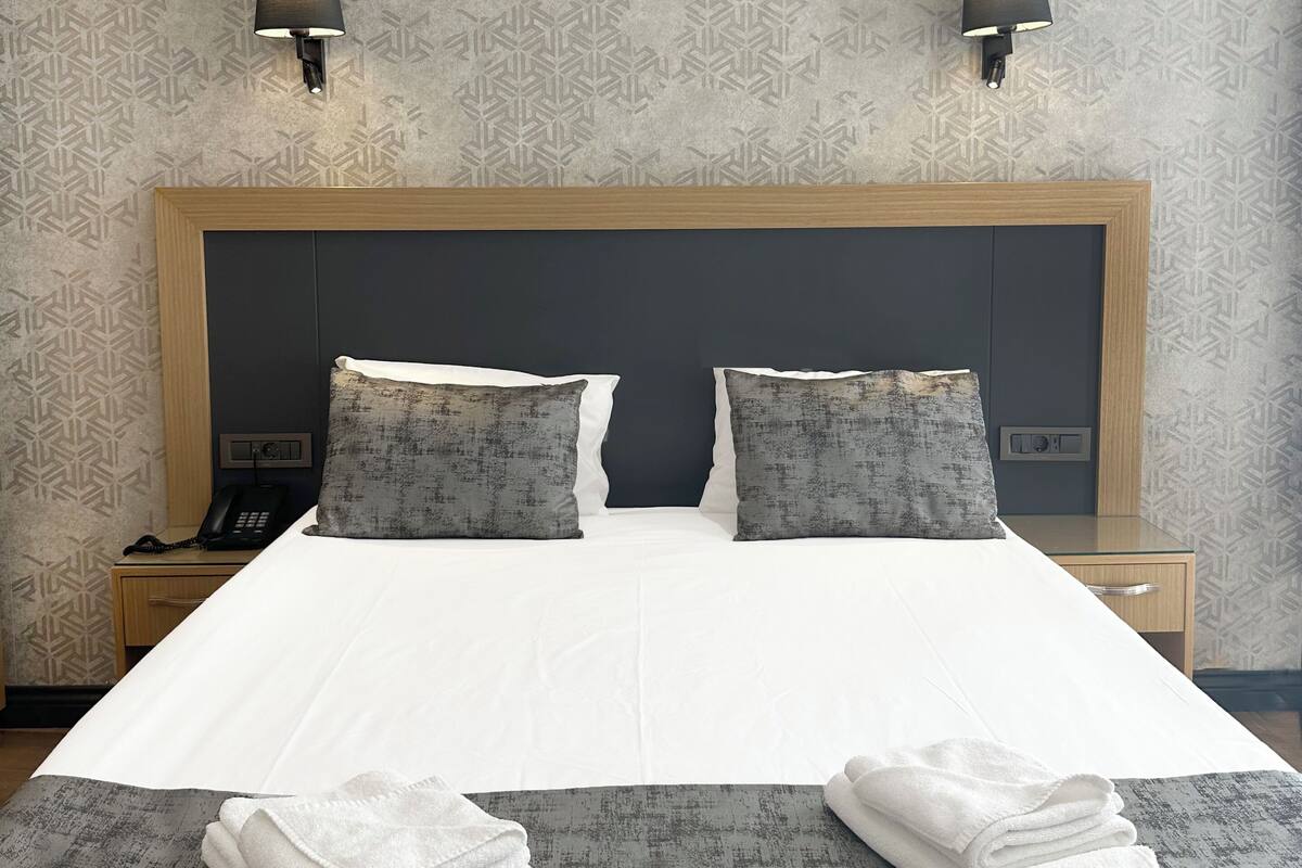 A hotel room with a large bed, two bedside lamps, a bedside phone, and a neatly folded towel on the bed.