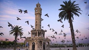 A historic tower with a clock, surrounded by palm trees and a plaza with birds flying around. - In Vivo Hotel (Izmir)