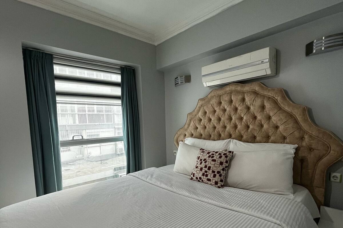 Una camera d'albergo con un letto grande, una finestra con tende, un condizionatore d'aria e un comodino.