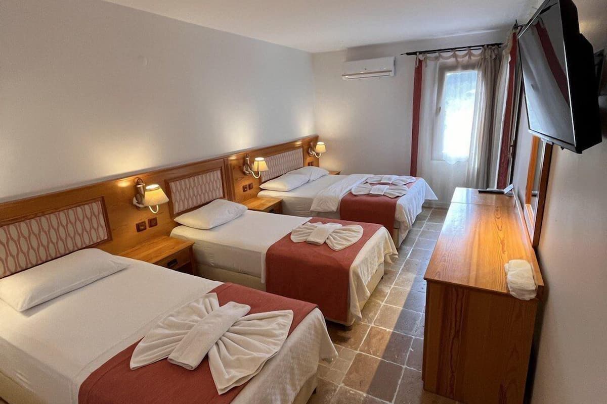 A hotel room with three single beds, each with a red bedspread and white towels. There is a wooden nightstand with a lamp, a flat-screen TV mounted on the wall, and a window with curtains.
