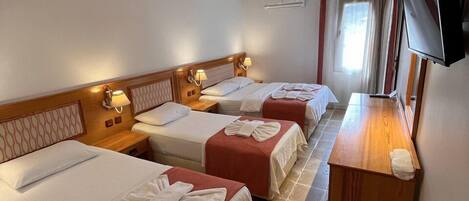 Habitación de hotel con tres camas individuales, cada una con colcha roja y toallas blancas. Se observa un mesita de noche de madera con lámpara, un televisor de pantalla plana en la pared y una ventana con cortinas.