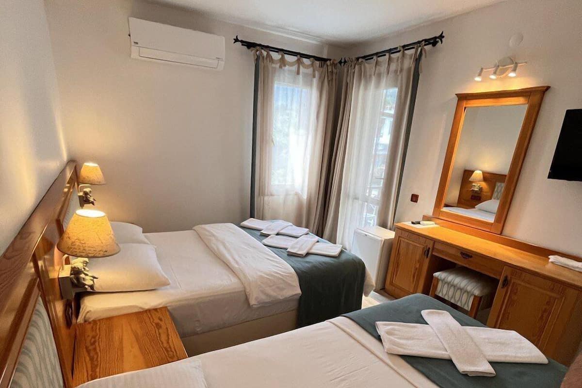 A hotel room with two beds, a wooden nightstand with a lamp, a small desk with a chair, a mirror, and a window with curtains.