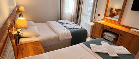 Habitación de hotel con dos camas, una mesita de noche con lámpara, un aparador de madera, un espejo y una ventana con cortinas.