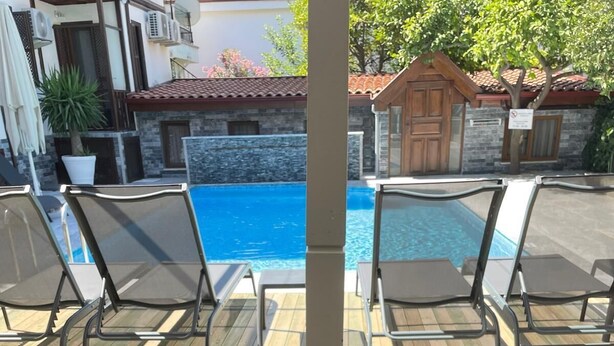A table with a variety of food items including bread, cheese, olives, and a drink, set against a backdrop of a pool and outdoor seating. - Yelken Hotel Akyaka (Ula)