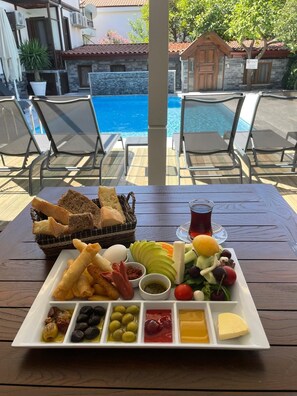 Una mesa con diversos alimentos, como pan, queso, aceitunas y una bebida, con una piscina y mobiliario exterior al fondo.