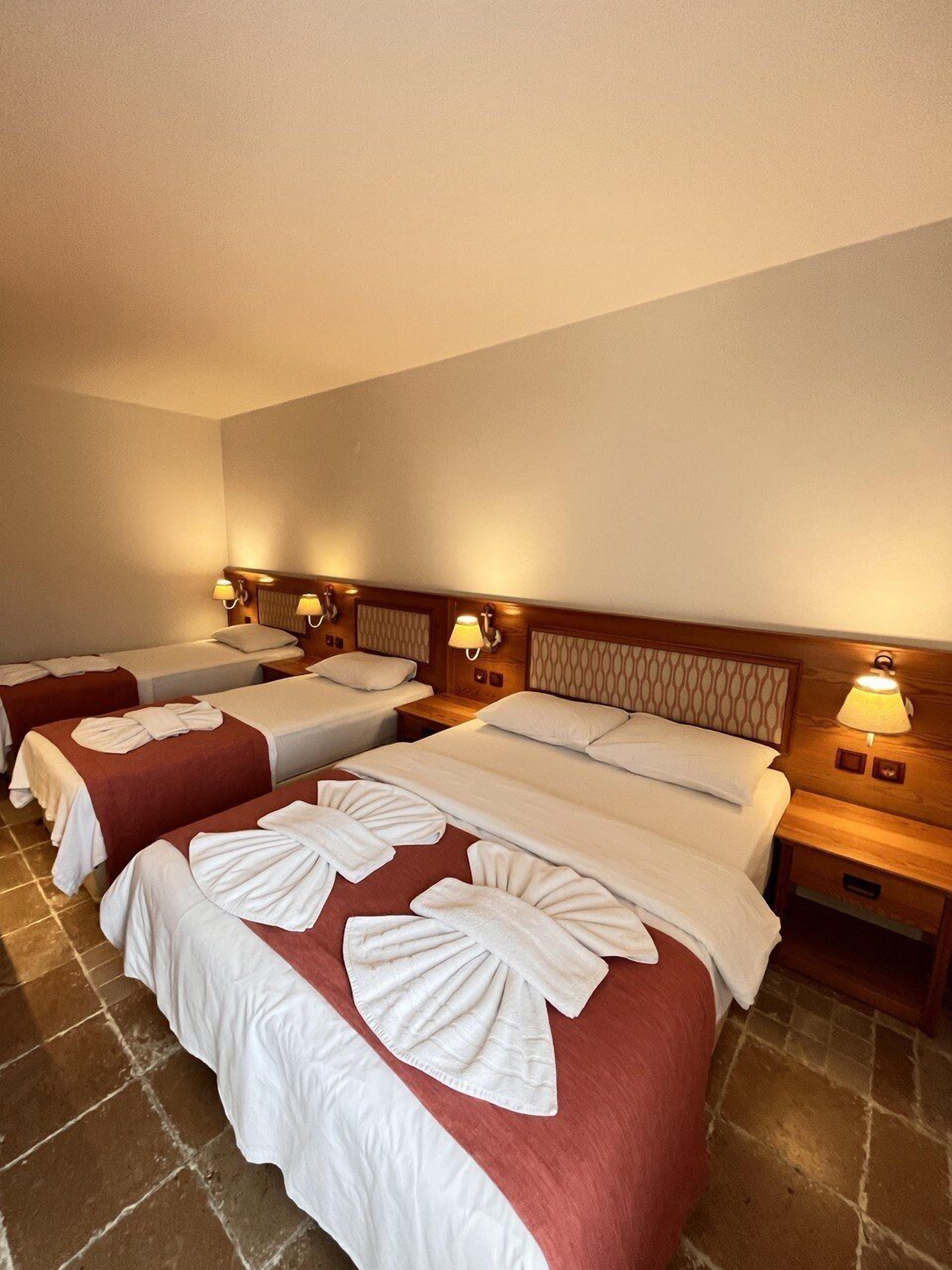 Habitación de hotel con tres camas, cada una con toallas impecablemente dobladas y una colcha roja. Se observan cabeceros y mesitas de noche de madera con lámparas.