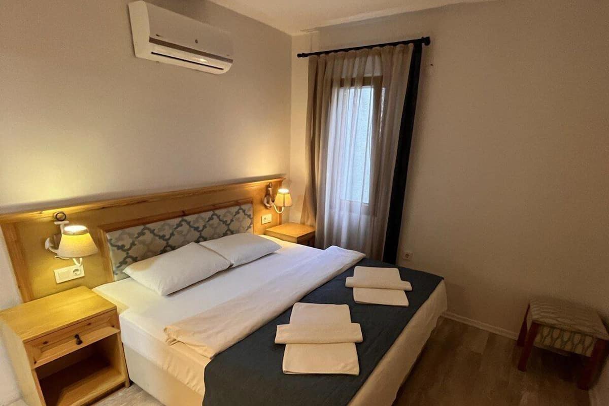 A hotel room with a bed, bedside tables, an air conditioning unit, and a window with curtains.