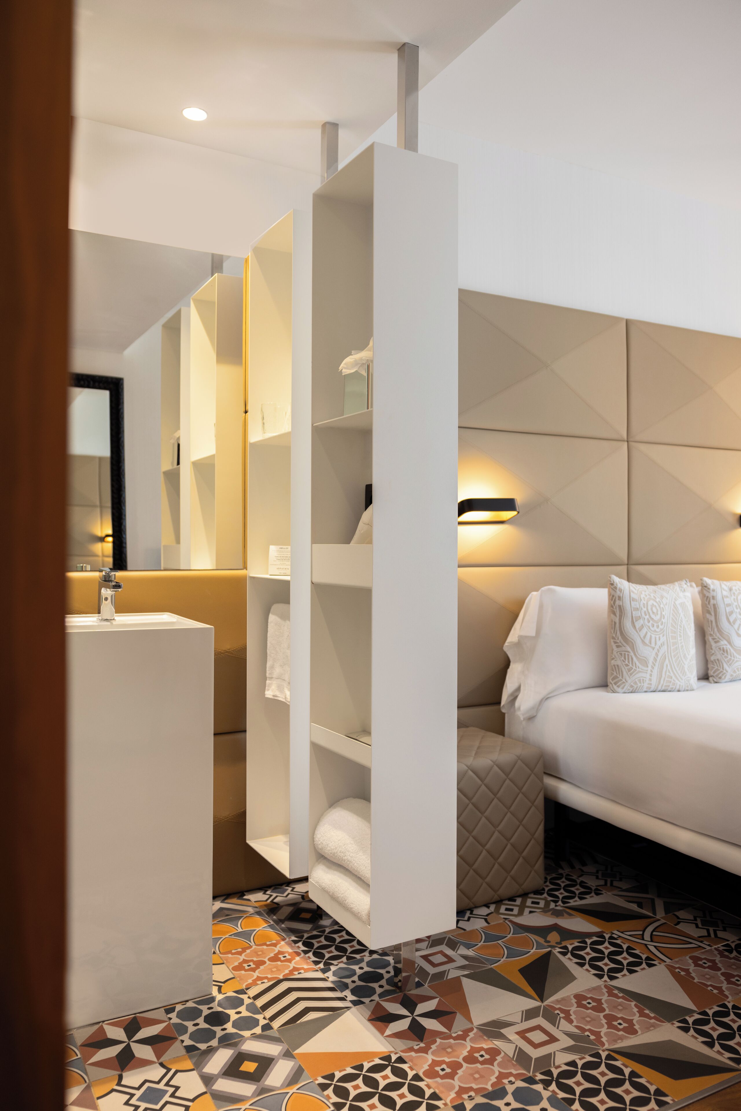 A modern bathroom with a white sink, a mirror, and a towel rack. There is a bed with a white headboard and patterned pillows.
