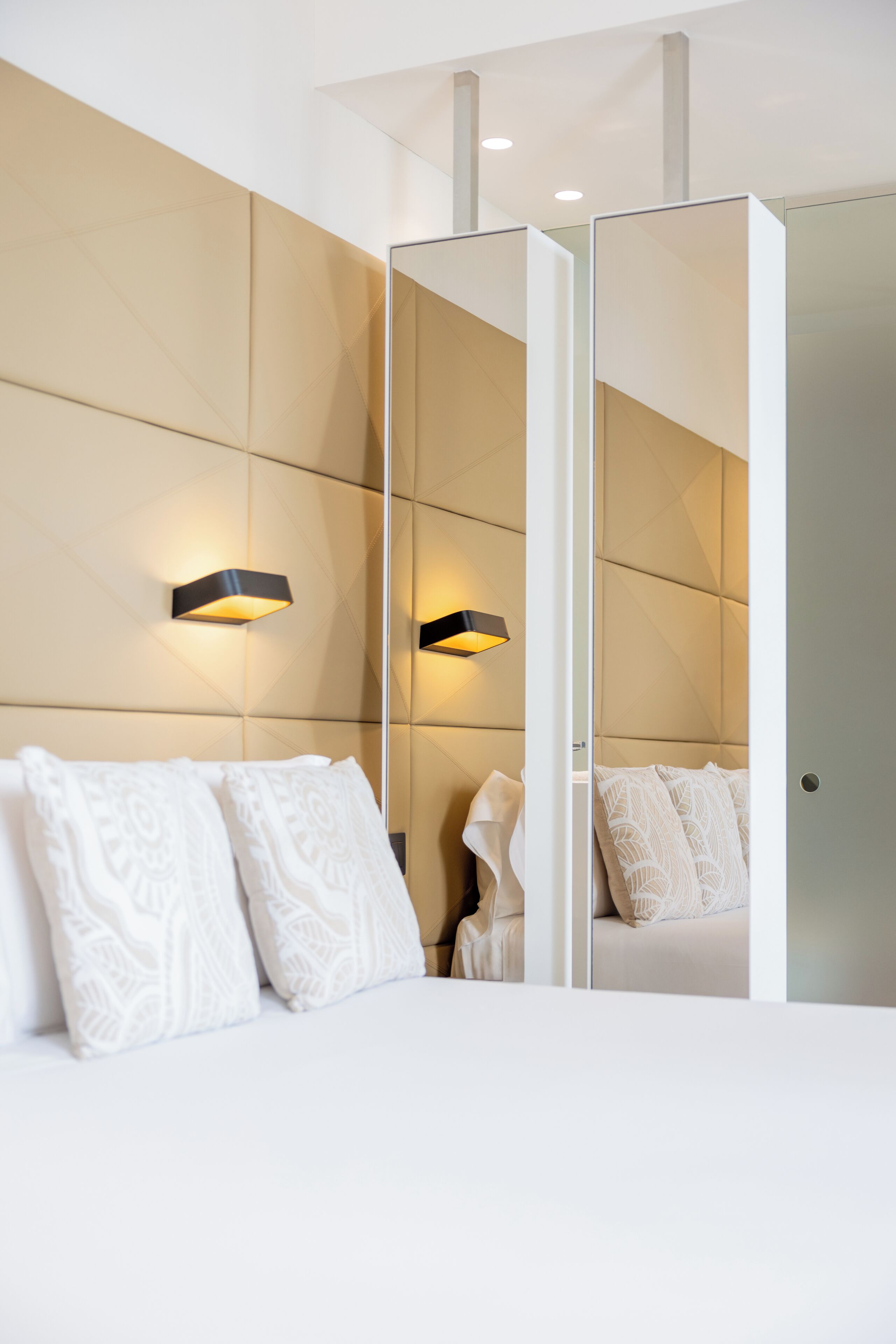 A modern hotel room with a bed, a large mirror, and wall-mounted lighting.