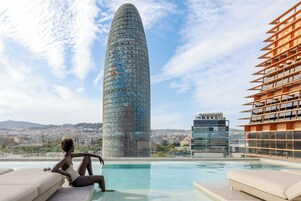 A modern building with a unique cylindrical shape, a rooftop pool area with lounge chairs, and a cityscape in the background.