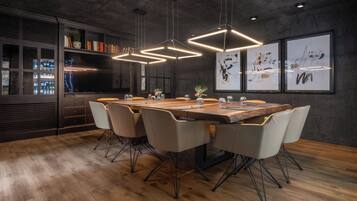 A modern dining area with a long wooden table, six chairs, pendant lights, and a bar area with shelves and a refrigerator.