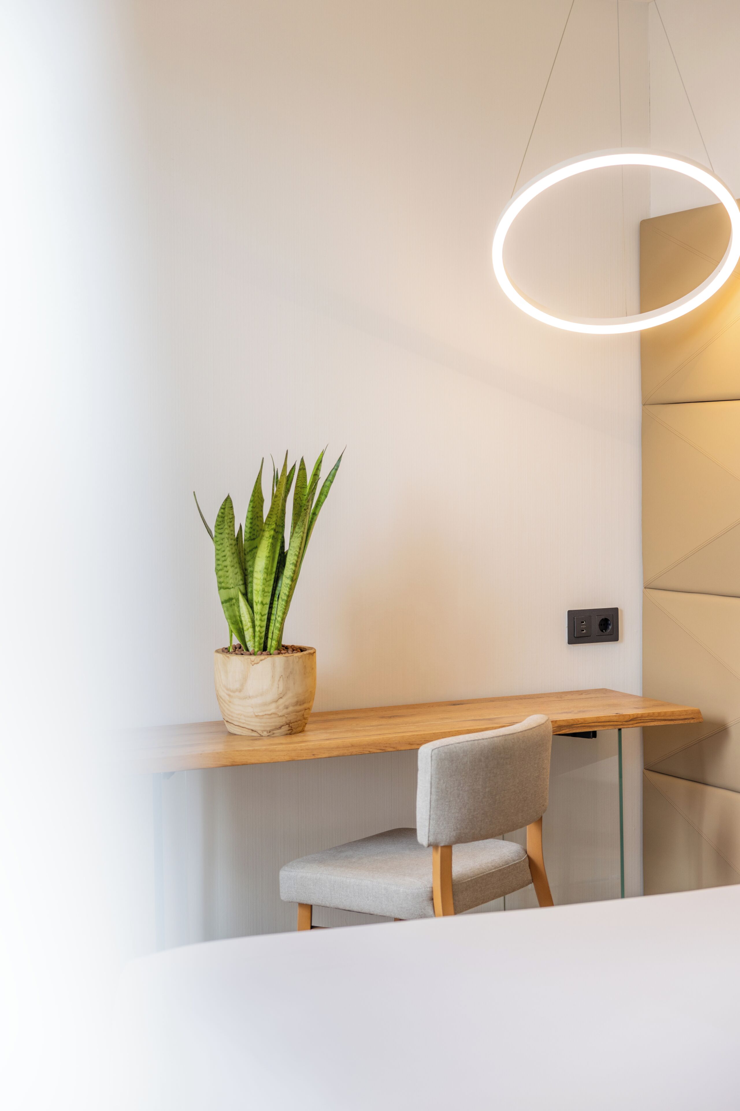 Una habitación moderna con un escritorio de madera, una silla gris, una planta en maceta y una lámpara circular.